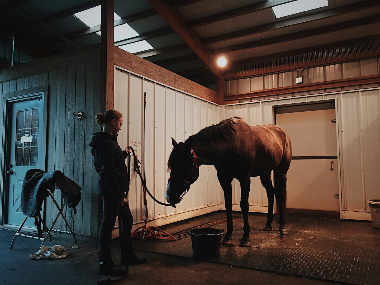 A yard owner leading a horse through a softly-lit barn aisle in the evening, saddle stand to the side.